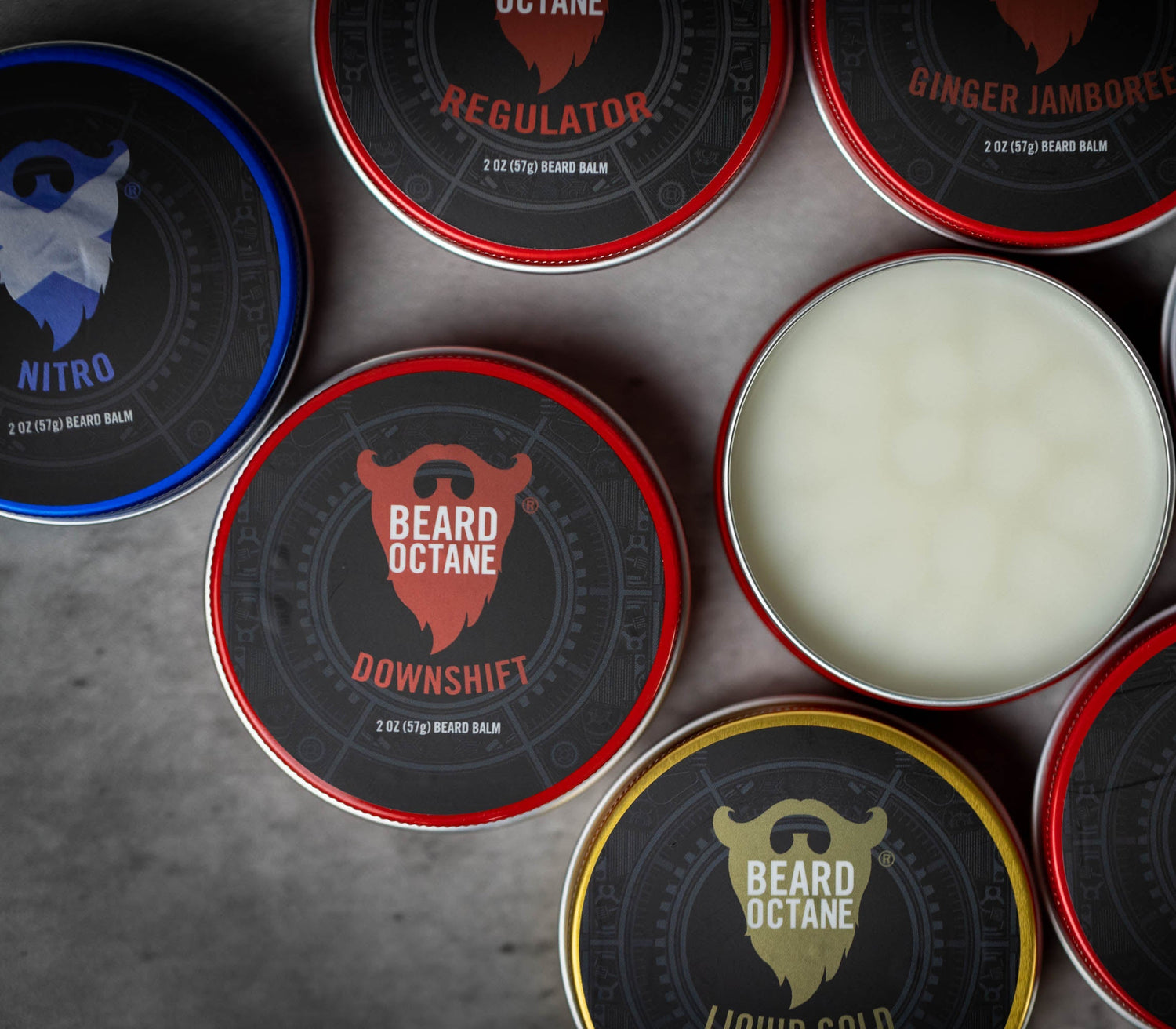 An overhead flat-lay of multiple round metal tins of Beard Octane beard balm, with one tin open to show the white balm inside. The tins feature different scent/variant labels including Nitro, Regulator, Ginger Jambonee, Downshift, Merica, Neutral, and Liquid Gold, arranged on a textured gray surface.