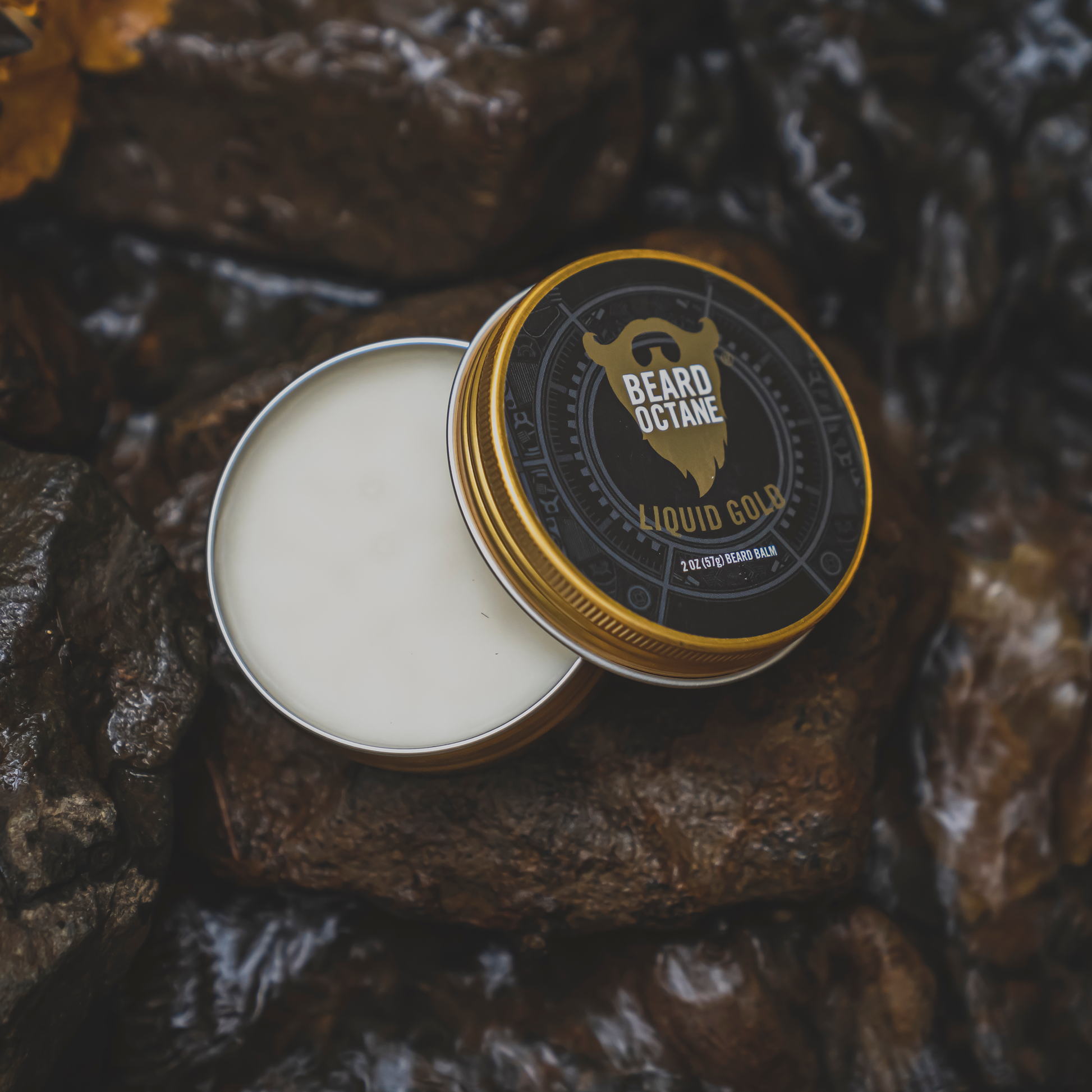 Close-up of a round tin of Liquid Gold Beard Balm with black and gold label featuring a beard icon, resting on dark, textured rocks